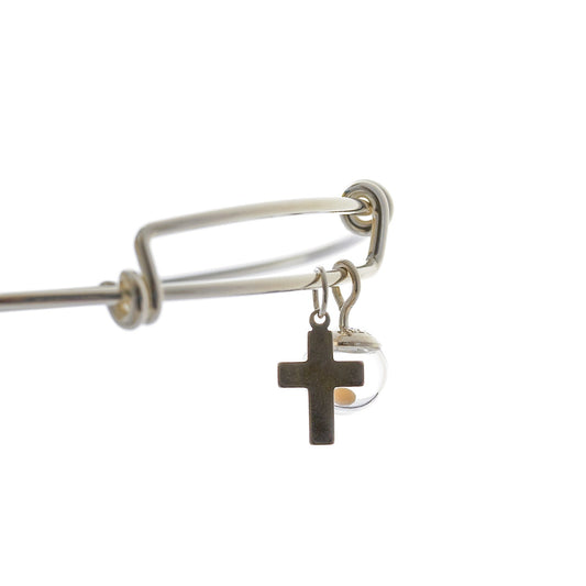 Close up of a sterling silver bangle bracelet featuring two dangling charms—a small mustard seed charm featuring a clear hand-blown glass globe containing a real mustard seed sealed with a sterling silver cap, and a simple sterling silver cross charm. The globe measures approximately 8mm and the charms hang freely from the sleek, adjustable bangle with coiled ends.