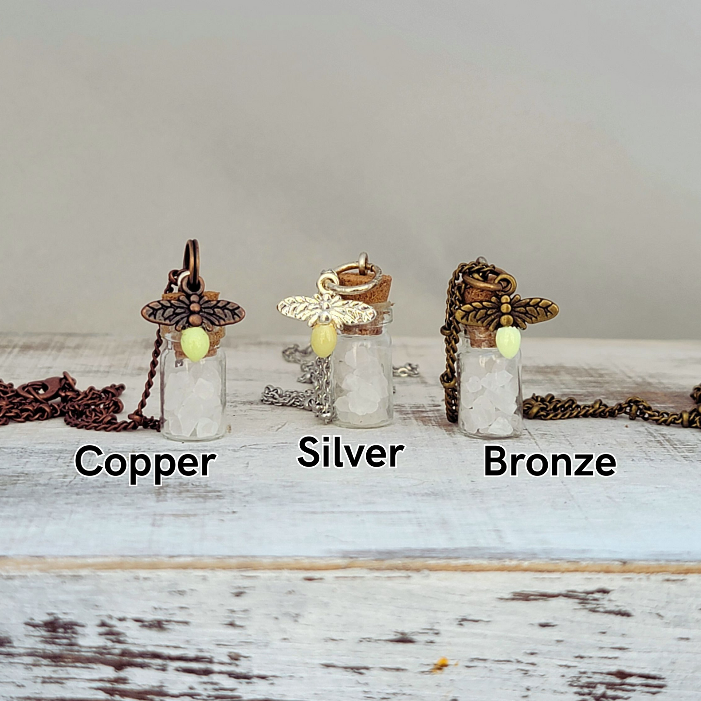 Photo of three small glass vial necklaces labeled Copper, Silver, and Bronze, each filled with coarse white salt crystals. Each vial is topped with a cork stopper and adorned with a decorative metal firefly charm witha small glow-in-the-dark accent designed to resemble a firefly’s light. The chains and metal components vary in color—copper, silver, and antique bronze—matching their respective labels. Displayed on a rustic whitewashed wooden surface with a soft gray background.