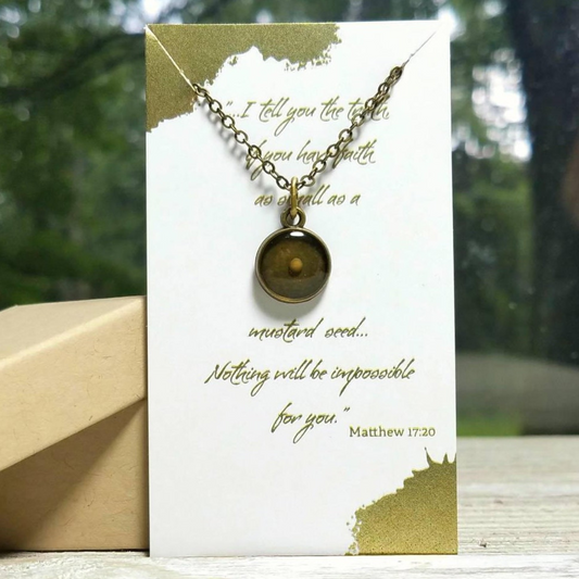A bronze mustard seed necklace with a glass-like clear resin bezel encasing a real mustard seed, presented on a card with gold foil-stamped text and a logo.