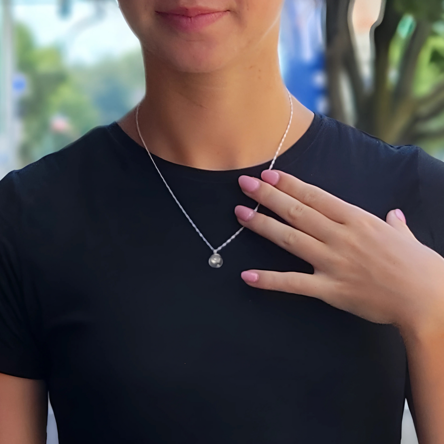 Woman wearing a sterling silver mustard seed necklace with a delicate Singapore chain, shown against a black top. She gently touches the pendant with her hand, highlighting the clear glass globe and tiny mustard seed inside. The background is softly blurred, focusing attention on the jewelry.