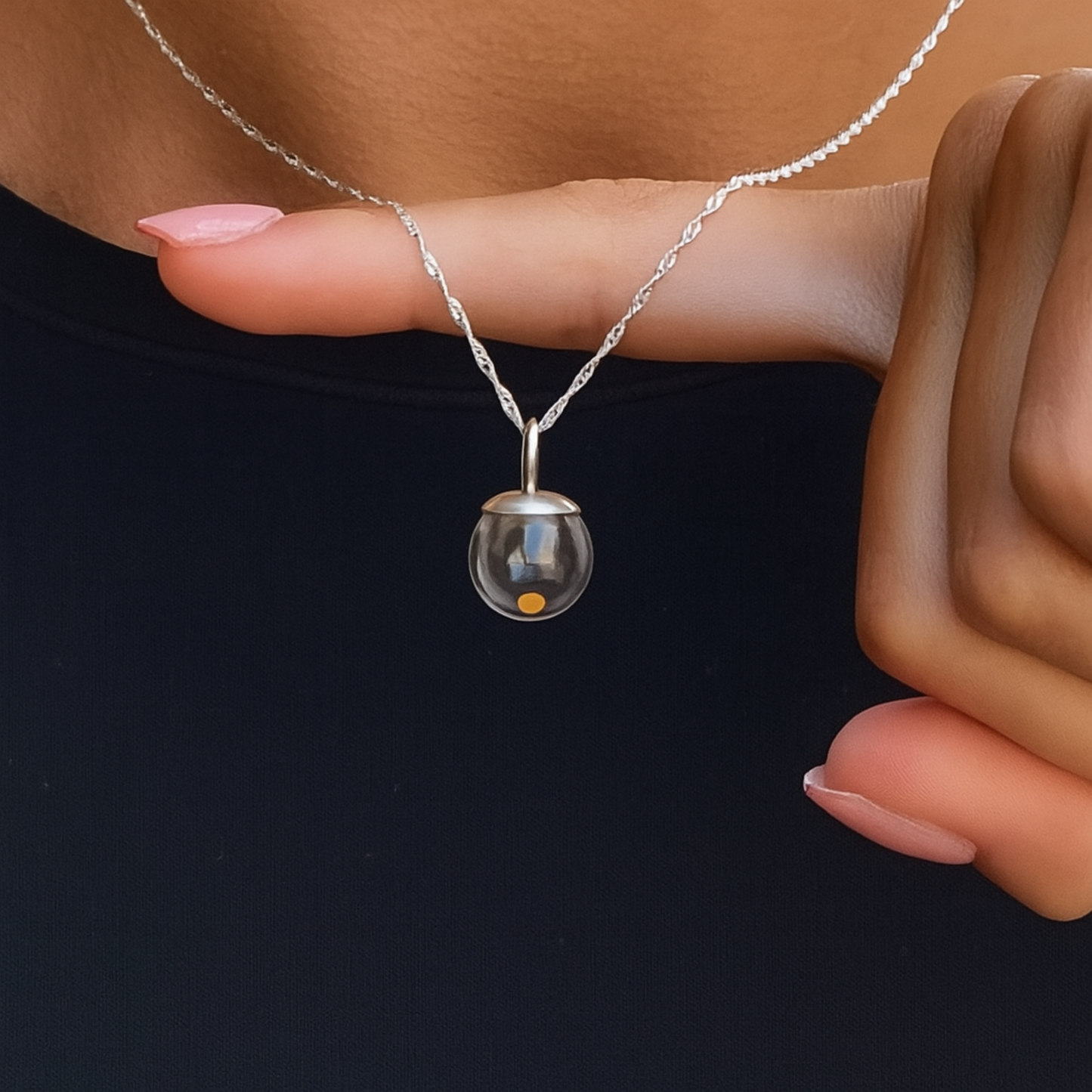 Close-up of a sterling silver mustard seed necklace with a delicate Singapore chain, hanging over a finger. The pendant features a tiny mustard seed encased in a clear glass globe with a silver cap, necklace is being worn against a black shirt for contrast.