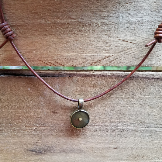 An antiqued bronze mustard seed pendant on a brown, adjustable distressed leather cord necklace laid out on a wooden surface.