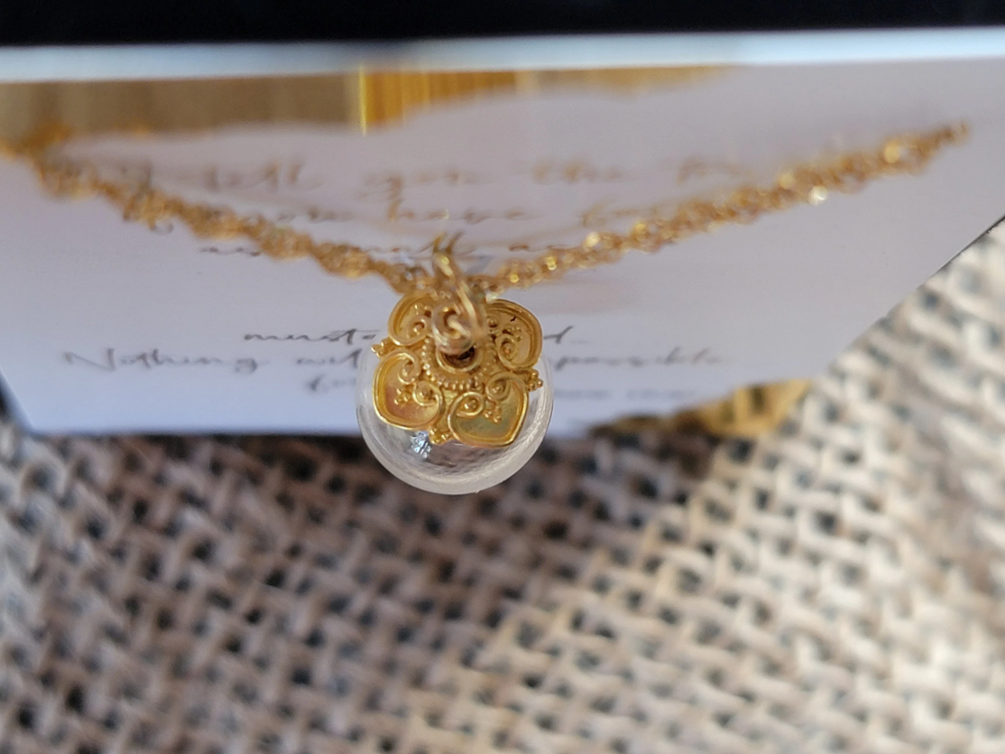 Close-up image of a gold mustard seed necklace featuring a delicate glass globe pendant with a real mustard seed that moves freely inside. The pendant is topped with an ornate, filigree-style solid gold cap and hangs from a fine 14k gold chain with a unique double loop wire wrapped gold bail. Behind the necklace is a scripture card partially visible with elegant gold script, resting on a woven burlap background.