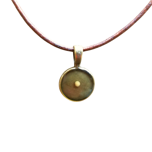 Round mustard seed pendant with a bronze bezel, featuring a single mustard seed encased in clear resin, hanging from a rustic brown leather cord necklace on white background.