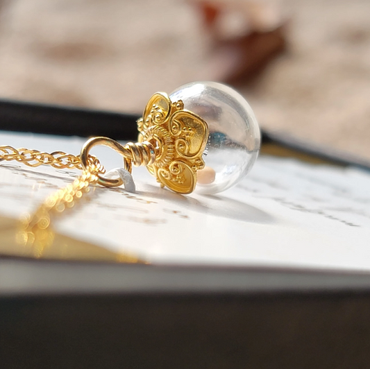 Close-up side view of a fancy gold mustard seed necklace featuring a clear hand-blown glass globe pendant with a single real mustard seed inside that moves freely. The pendant is topped with an ornate solid gold filigree cap with a wrapped gold bail, which connects it to a beautiful gold chain. The necklace rests on a partially visible scripture card, with a softly blurred background adding warmth and depth.