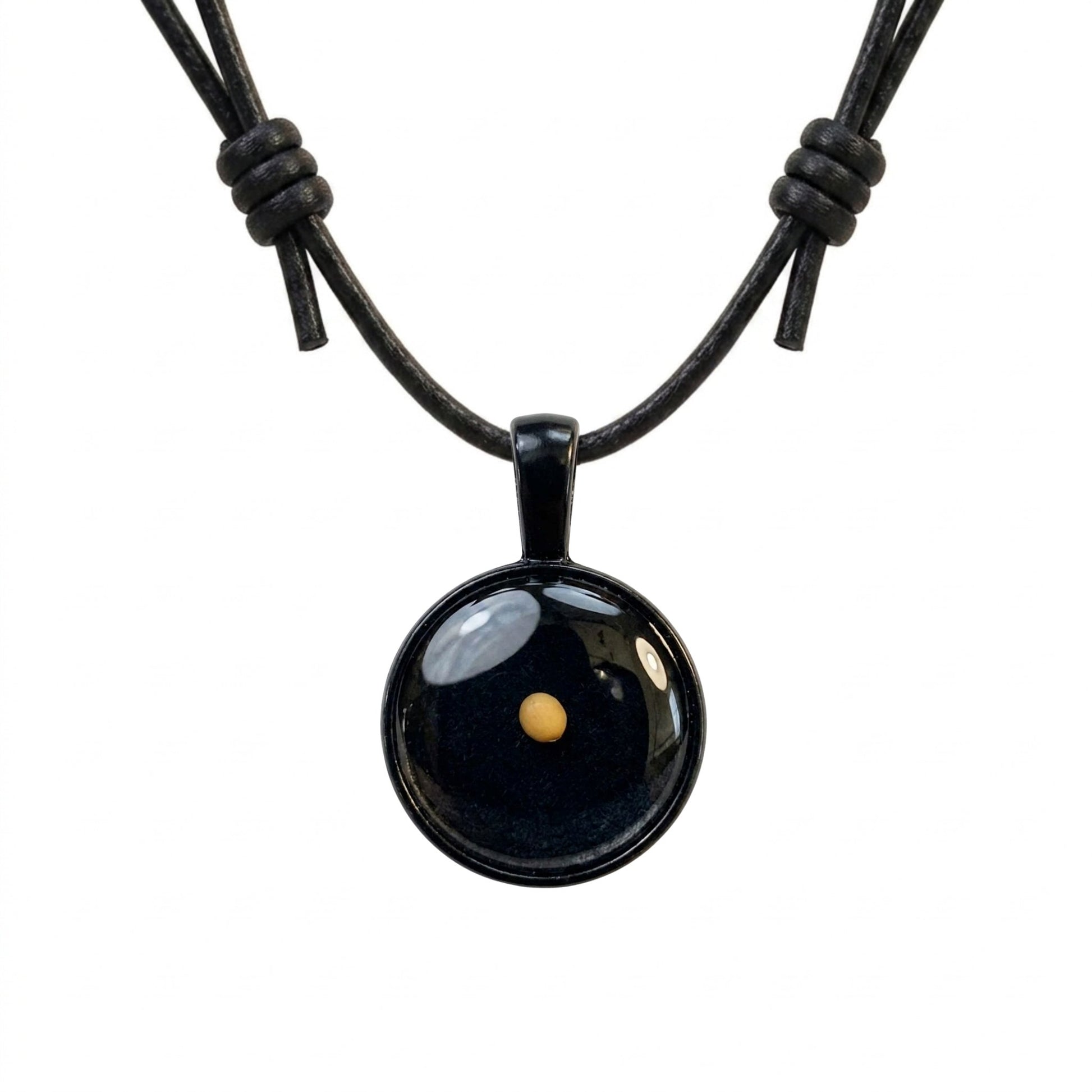 A product image of a single mustard seed encased under a clear resin dome pendant with a gunmetal black bezel housing, hanging from an adjustable black leather cord necklace with decorative barrel knots on a pure white background.