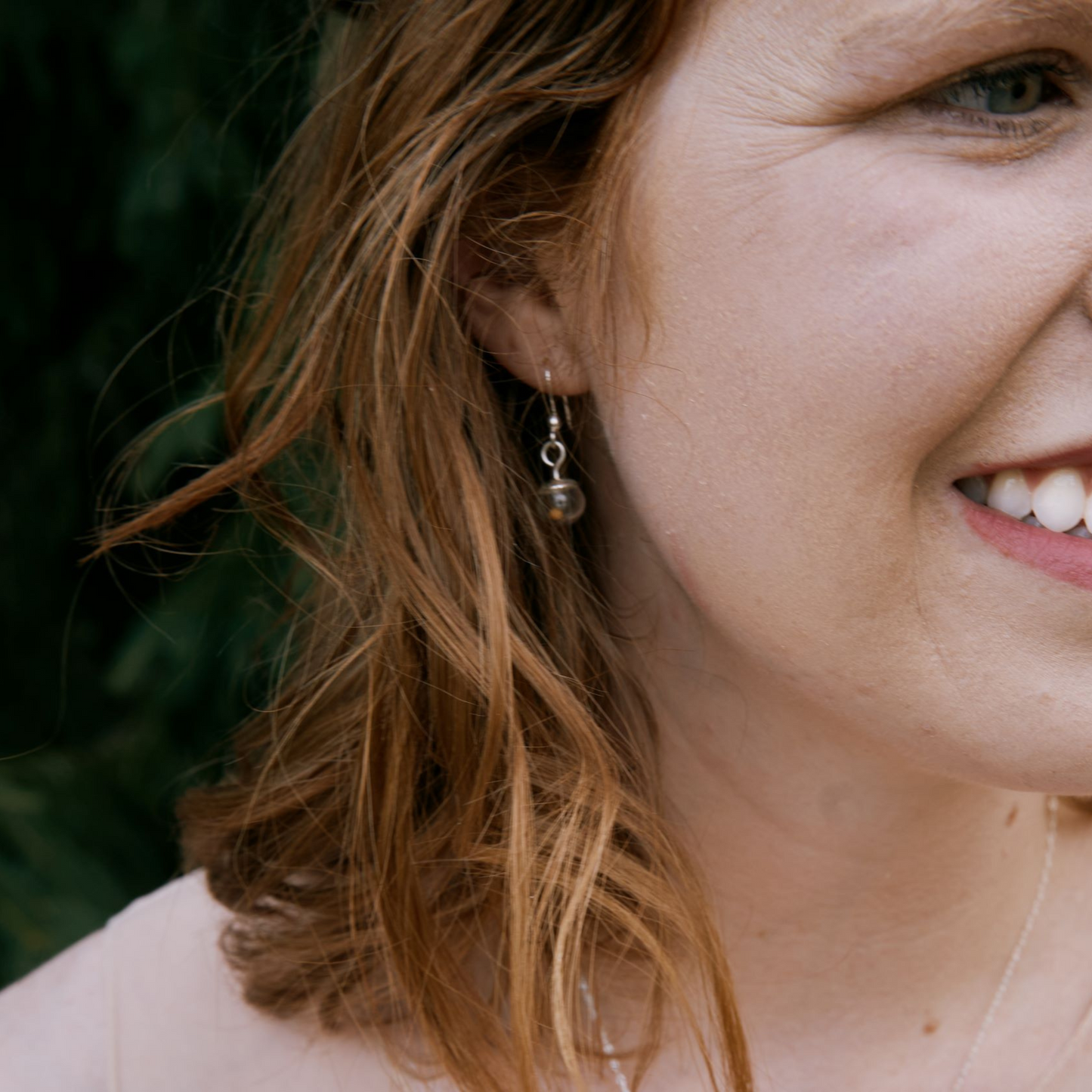 Close-up of a smiling woman with wavy auburn hair wearing a dangling earring made from a small glass globe containing a mustard seed, dangling from a sterling silver shepherd’s hook.