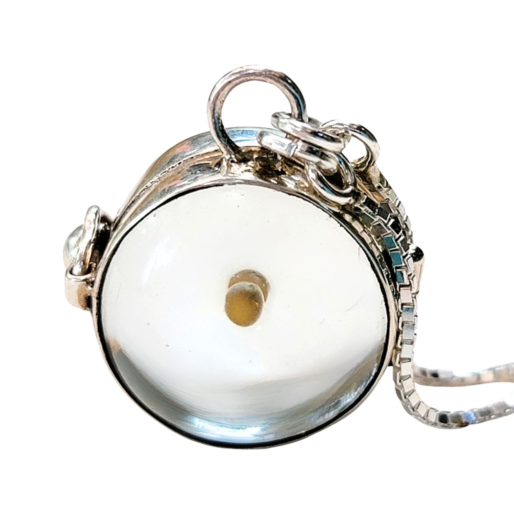 Close-up image of a round sterling silver mustard seed pendant featuring a single real mustard seed sealed inside a clear, domed glass globe. The pendant is set in a smooth polished sterling silver bezel and includes a functional hinge and clasp. A silver loop with two jump rings connect the pendant to a sleek, sterling adjustable box chain.