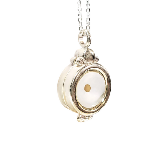 A close-up image of a 925 sterling silver pendant necklace featuring a round magnetic locket with glass & resin centerpiece with a small real mustard seed sealed in the middle with clear resin that can be seen from both the front and the back. The pendant has a slightly industrial design with decorative metalwork and is suspended from a delicate beveled sterling silver cable chain.