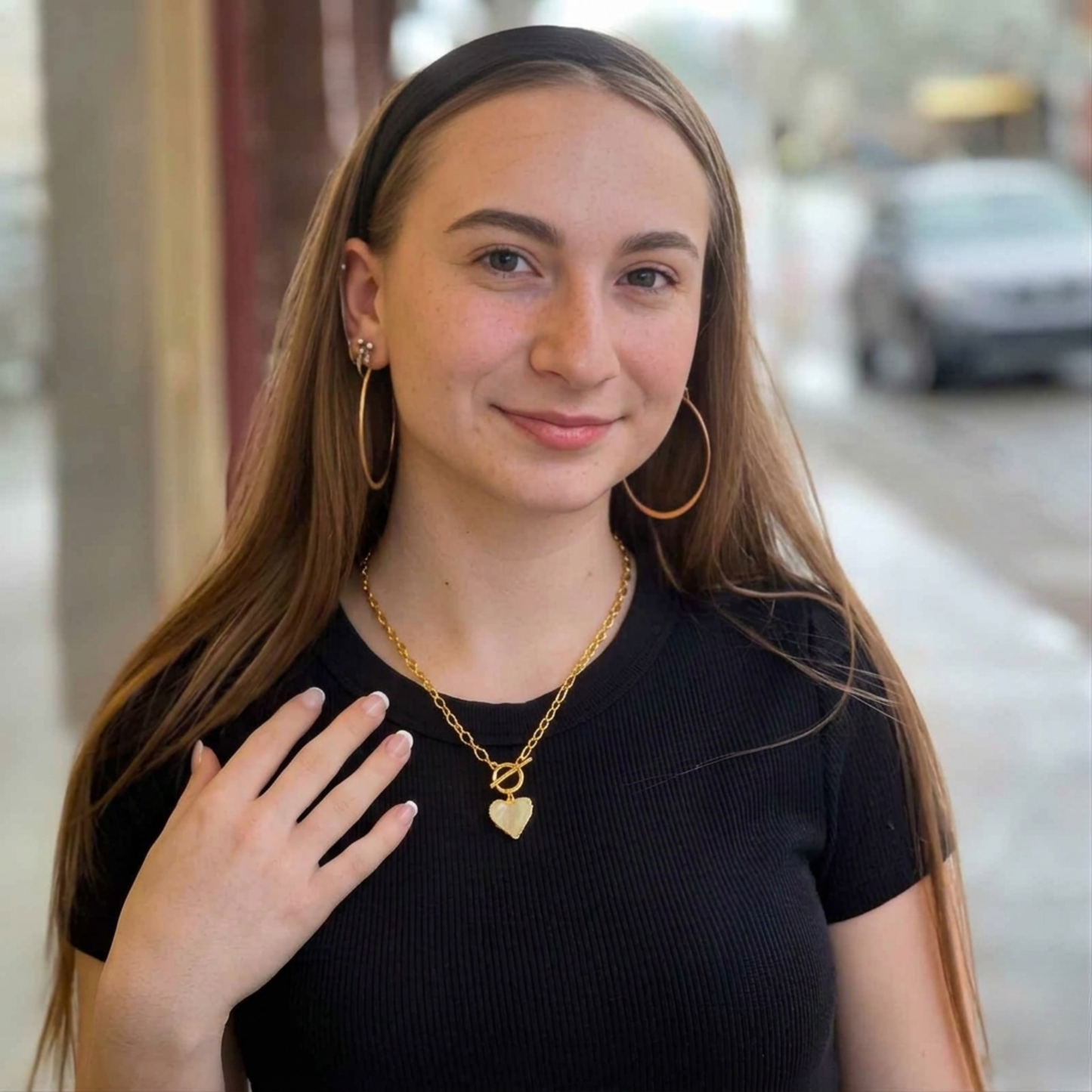 A lifestyle photo of a young woman smiling outdoors, wearing a gold paperclip-link necklace with a heart-shaped pendant. She is dressed in a black top and a black headband, accessorized with large gold hoop earrings. The necklace features a front-facing toggle clasp that holds a light-colored, textured heart pendant. The background is a softly blurred street scene, highlighting the jewelry as a stylish, everyday accessory.