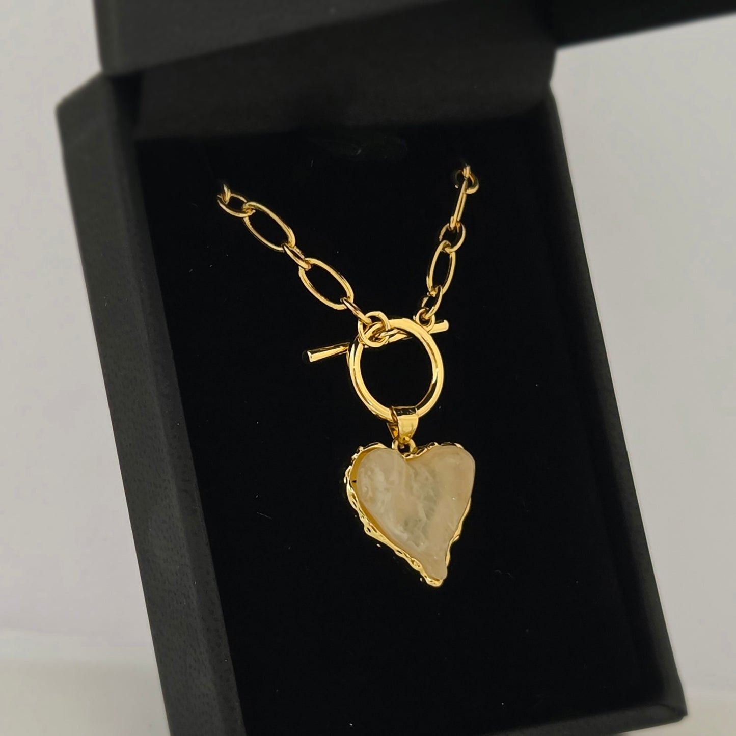 A gold paperclip-link necklace with a toggle clasp is presented in an open black jewelry box. Hanging from the clasp is a heart-shaped pendant with a textured, light milky pink colored acrylic stone center and a decorative gold-tone border. The jewelry rests against a dark velvet interior, highlighting the bright gold finish and the unique, organic shape of the heart pendant.
