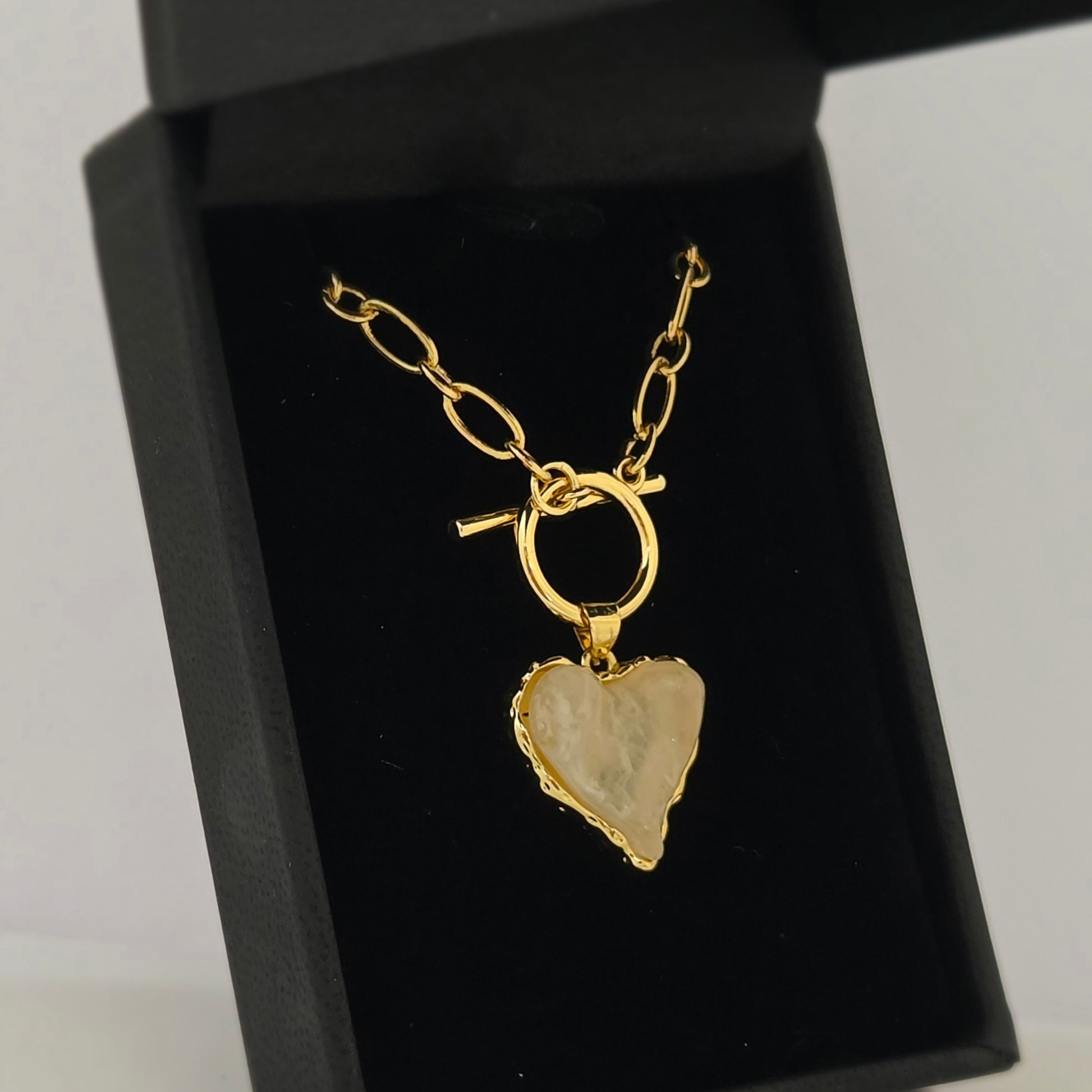 A gold paperclip-link necklace with a toggle clasp is presented in an open black jewelry box. Hanging from the clasp is a heart-shaped pendant with a textured, light milky pink colored acrylic stone center and a decorative gold-tone border. The jewelry rests against a dark velvet interior, highlighting the bright gold finish and the unique, organic shape of the heart pendant.