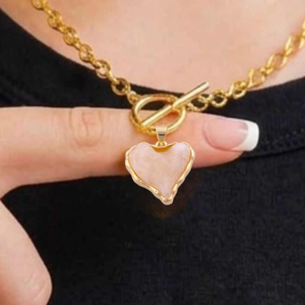 A close-up lifestyle shot shows a person wearing a gold-tone chain paperclip necklace with a front-facing toggle clasp. A small, light milky pink acrylic heart-shaped pendant with an organic, wavy gold border hangs from the clasp. The woman is wearing a black 