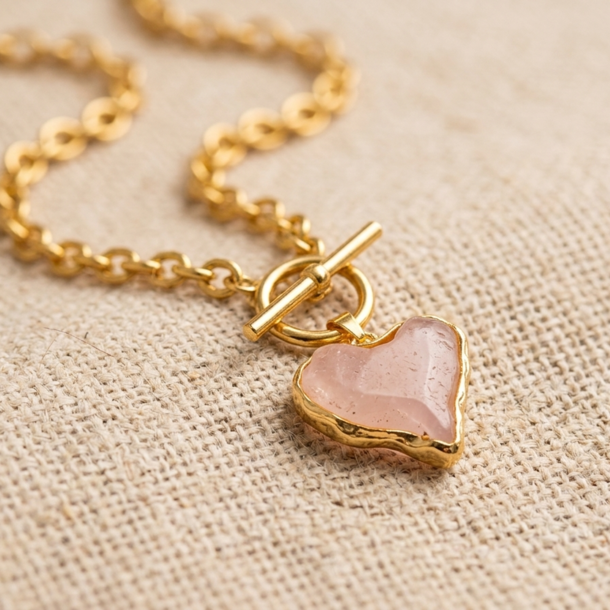 A close-up shot of a gold necklace with a paperclip-link chain and a front-facing toggle clasp. A light milky pink, heart-shaped acrylic pendant hangs from the toggle, featuring an organic, slightly irregular shape and a textured gold border. The jewelry is displayed on a neutral, textured linen fabric background, emphasizing the delicate color and elegant design.