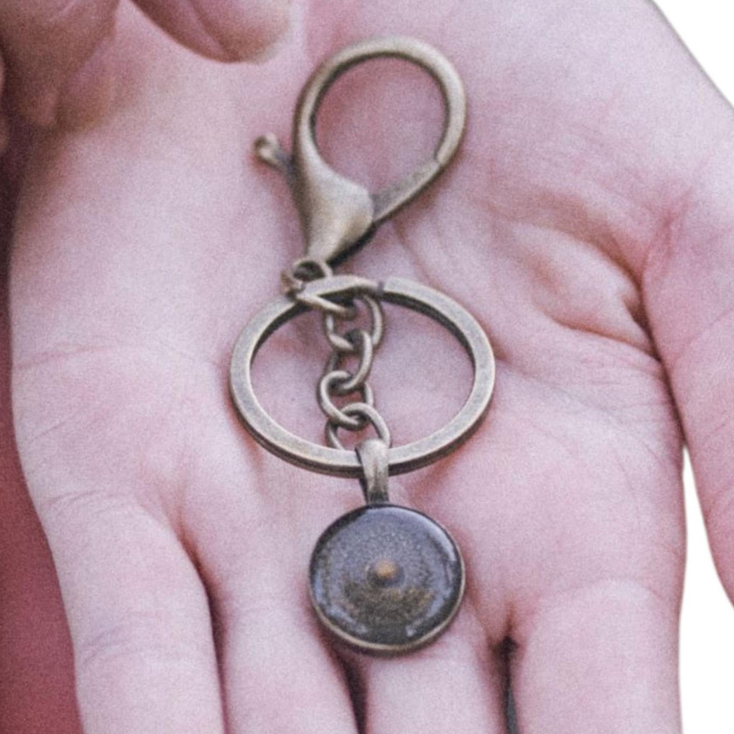 An antique bronze mustard seed keychain held in a person's hand, featuring a small circular bronze bezel with a real mustard seed in the center, attached to a keyring and a lobster-style clasp.