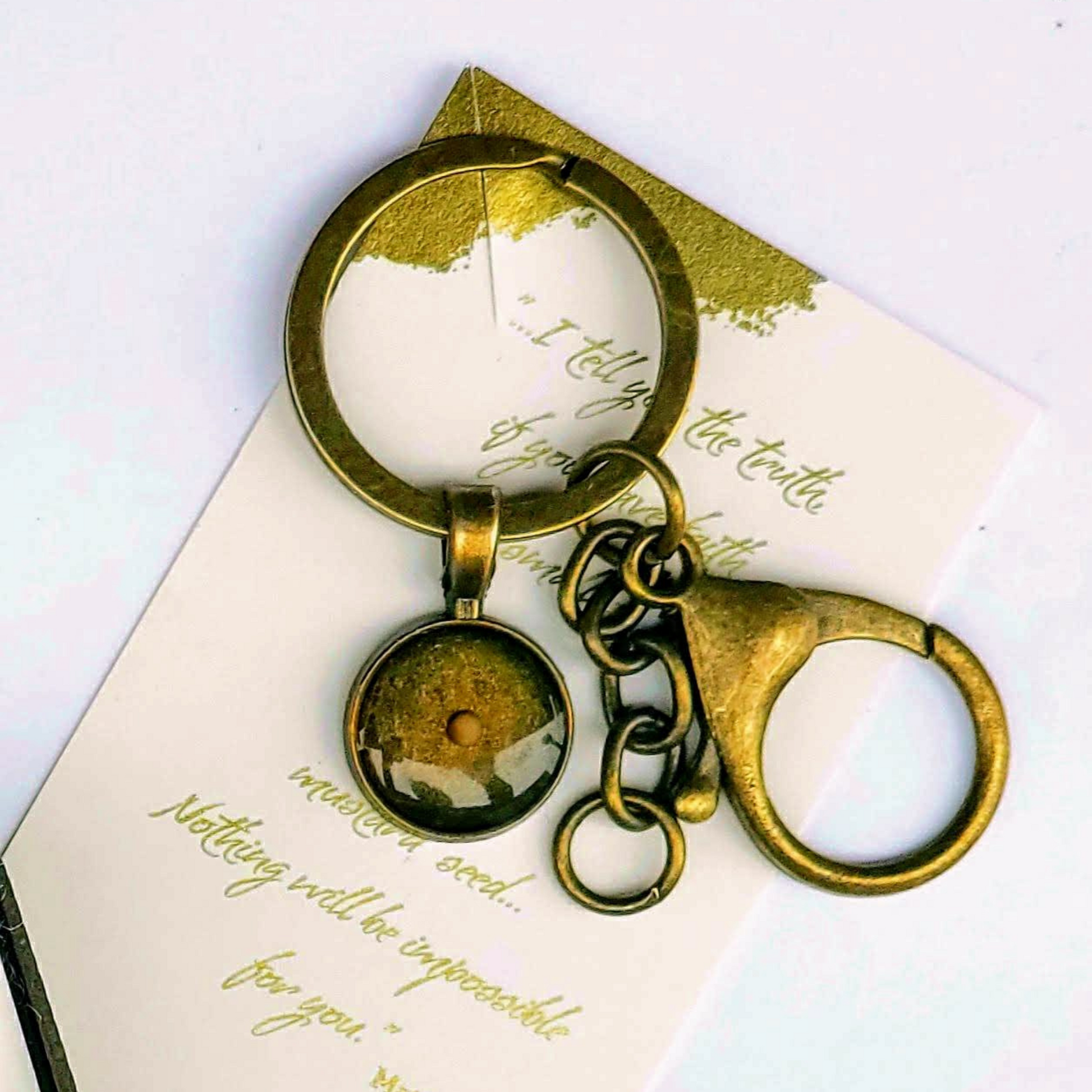 Small bronze mustard seed keychain with a round resin pendant encasing a real mustard seed, attached to a bronze keyring and large lobster claw clasp with chain links. The keychain is displayed on a decorative scripture card featuring Matthew 17:20, which reads, “I tell you the truth, if you have faith as small as a mustard seed… Nothing will be impossible for you,” with gold foil-style accents in the corners.